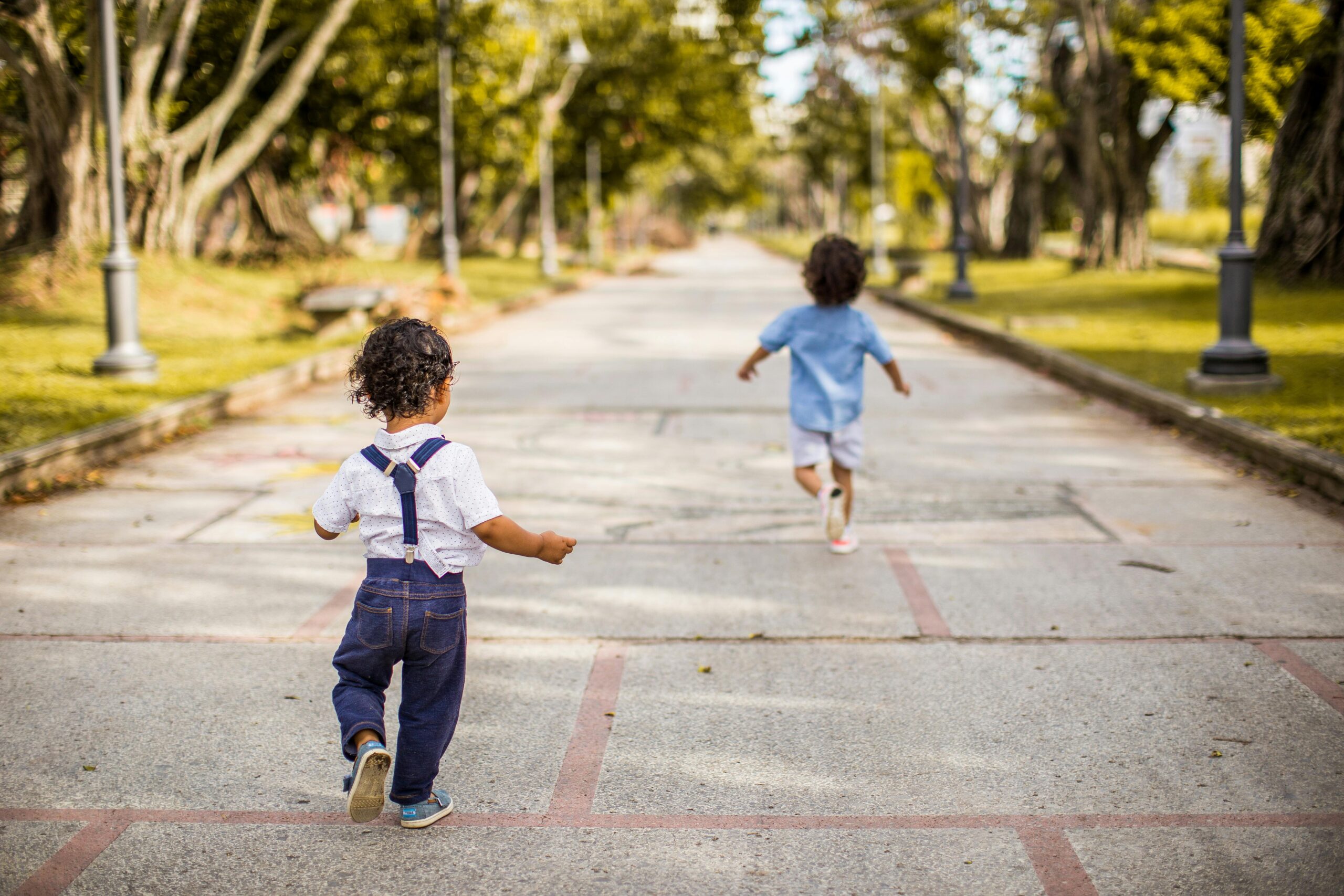 Two young children running joyfully on a paved pathway in a scenic park setting, enjoying a sunny day.